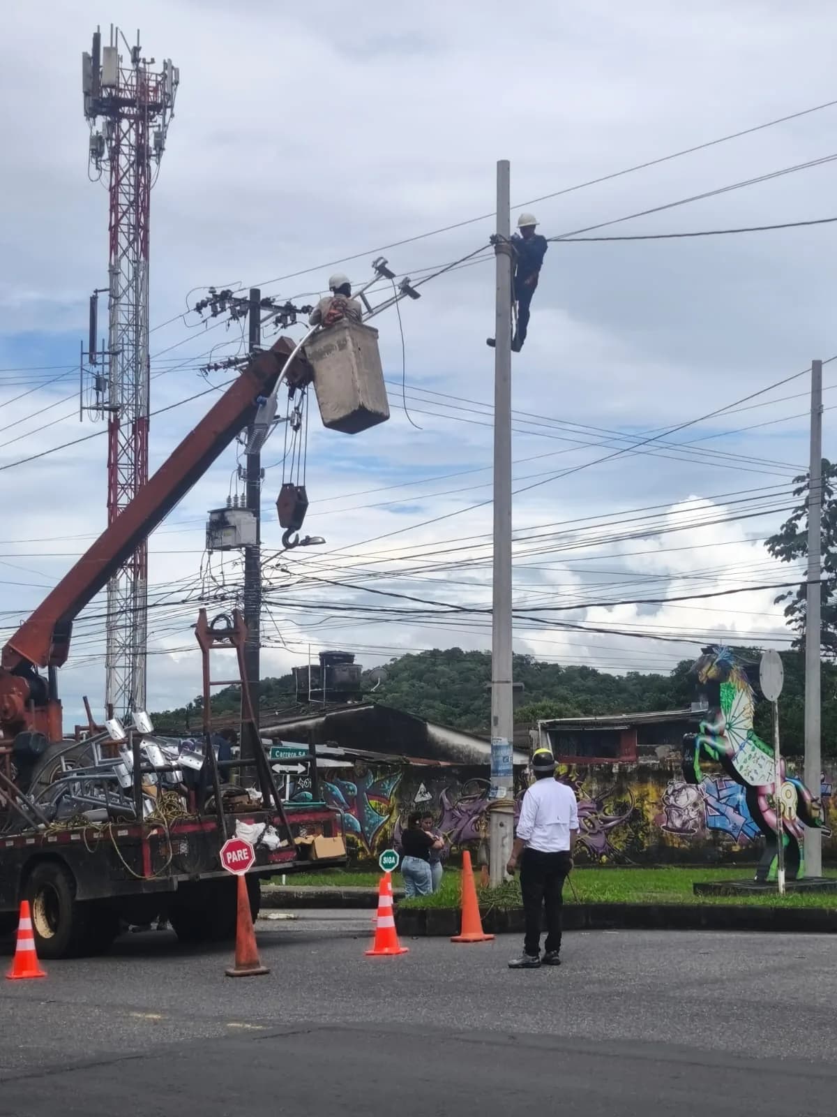 Camión grúa con canasta elevando a un técnico junto al poste mientras otro operario asciende con arnés; en el platón se ven las luminarias de sodio retiradas y la señalización vial de la obra.