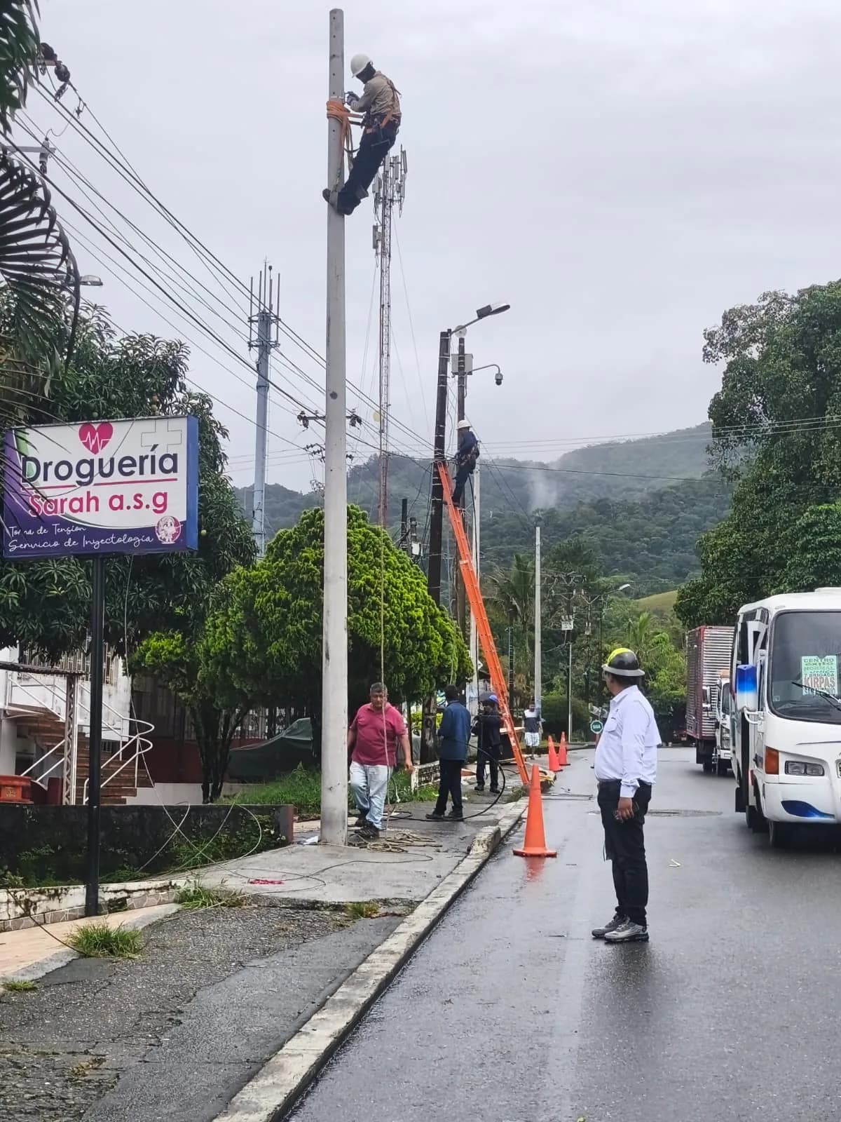Dos técnicos trabajando en altura sobre poste y escalera durante la jornada de lluvia, con conos de seguridad y el supervisor de obra de espaldas sobre la vía mojada.
