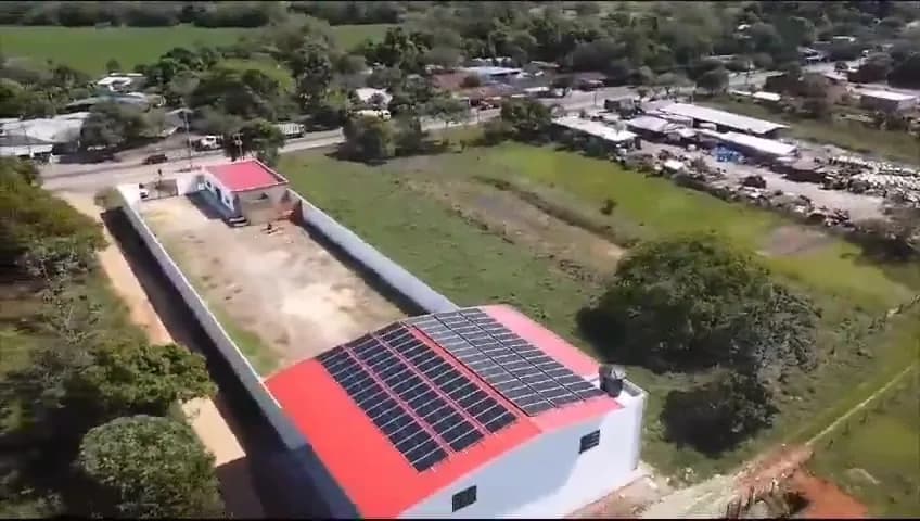 Vista con dron de la bodega terminada: la cubierta roja con el arreglo solar completo y el patio de volquetas y maquinaria del predio.