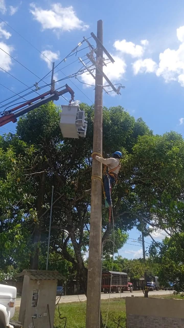 Camión grúa izando el transformador hasta el poste mientras un técnico asciende para recibirlo.