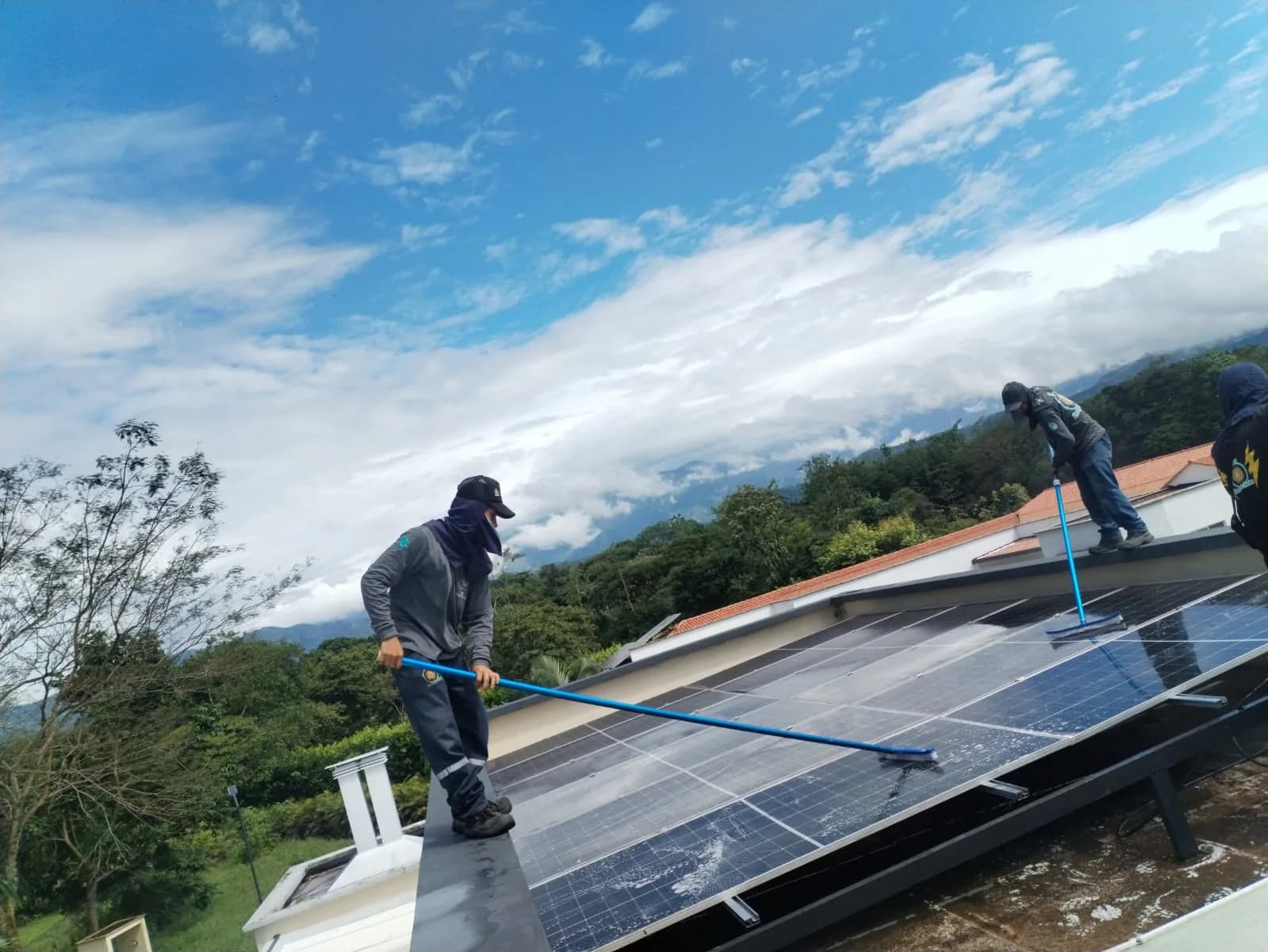 Dos técnicos de Voltaje Zero lavando con cepillo telescópico el arreglo de la azotea, con la cordillera y las nubes bajas de fondo.