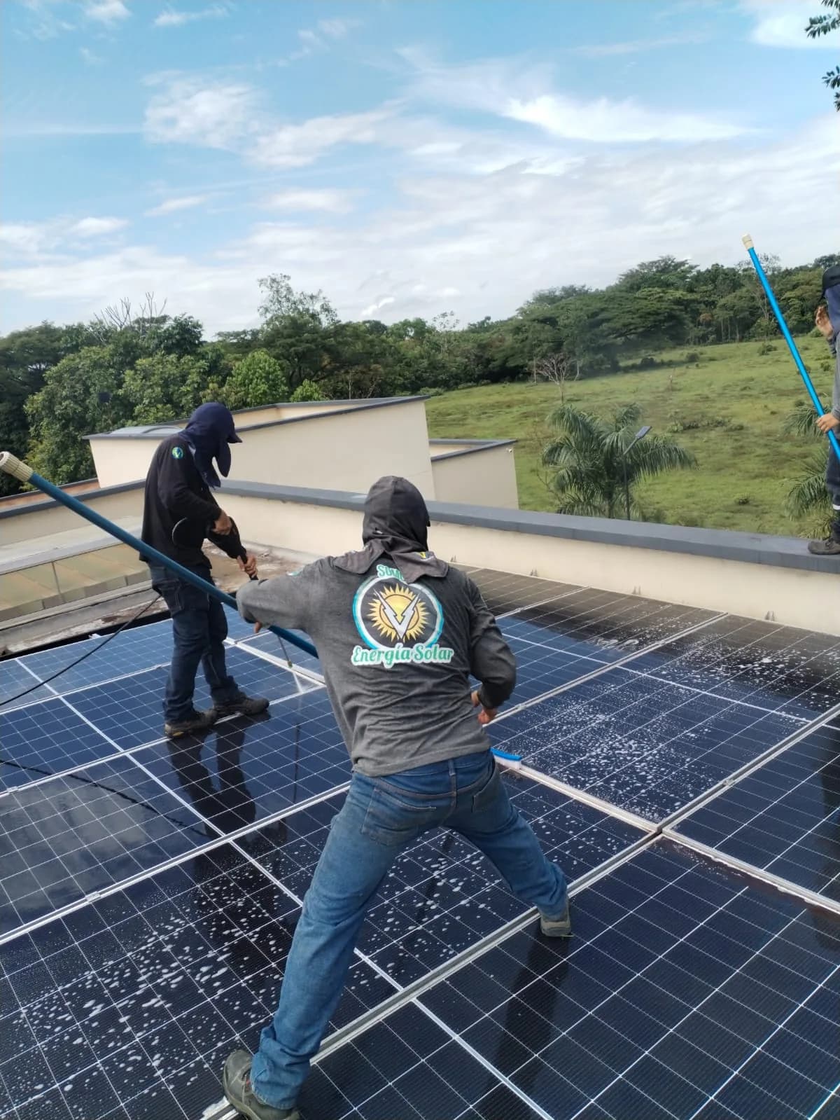 Cuadrilla de Voltaje Zero lavando con agua y cepillo los módulos de la azotea, de espaldas y con el uniforme de energía solar visible.