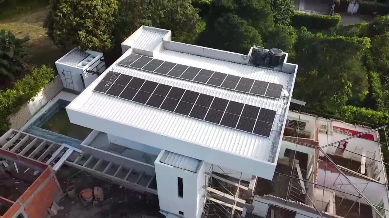 Vista aérea cercana de la cubierta metálica blanca de la casa campestre con las tres filas de módulos solares instaladas, junto a la piscina y la ampliación en obra.