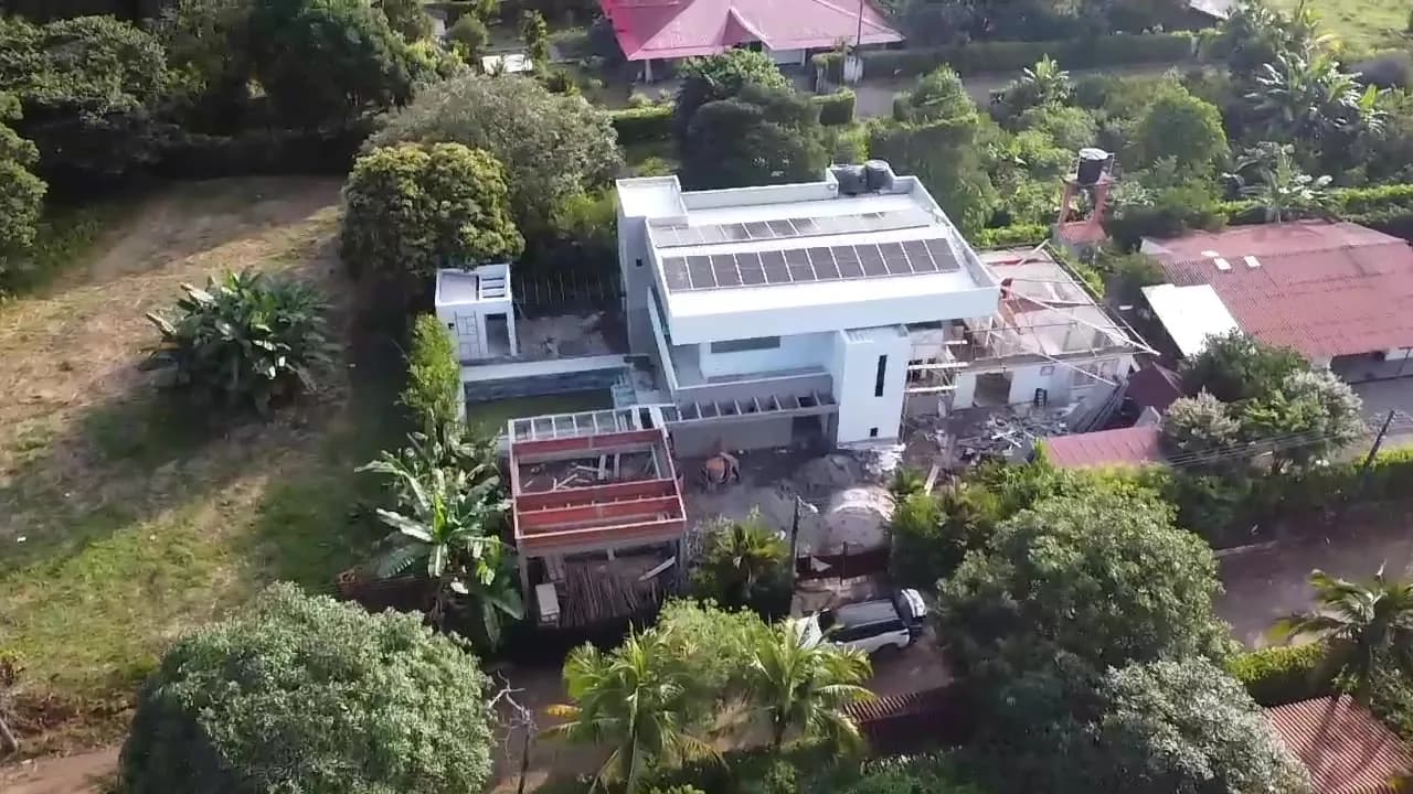 Panorámica aérea de la casa campestre completa con el arreglo solar en la cubierta, rodeada de jardines, palmas y las casas vecinas.