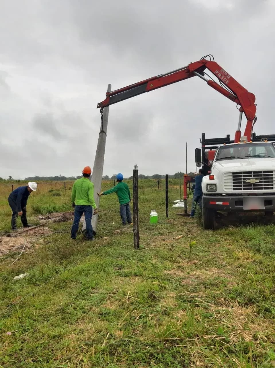 Poste de concreto izado en diagonal por el brazo hidráulico del camión grúa mientras la cuadrilla lo guía hacia la excavación.