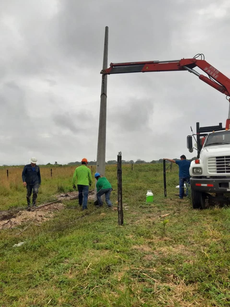 Poste ya vertical sostenido por la grúa mientras los operarios lo alinean sobre el hoyo, con el camión y el potrero al fondo.
