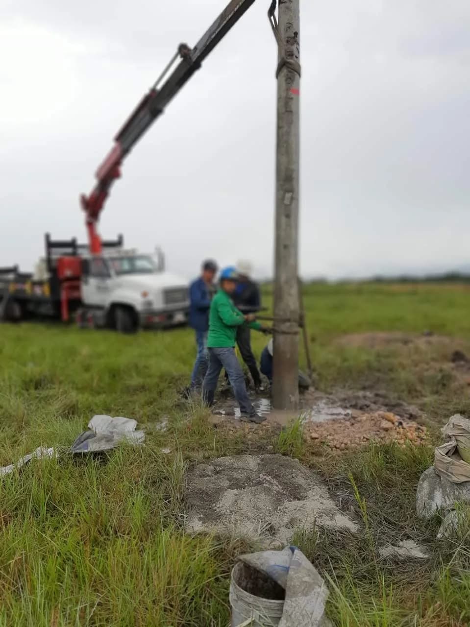 Detalle de la base del poste, con el agua de la excavación, los bultos de cemento y la cuadrilla trabajando al fondo.